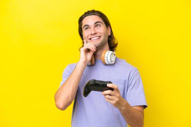 Young handsome caucasian man playing with a video game controller over isolated on yellow background thinking an idea while looking up