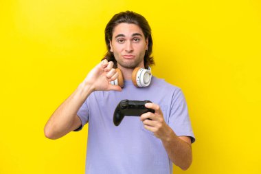 Sarı arka planda izole edilmiş bir bilgisayar oyunu kontrolörüyle oynayan yakışıklı beyaz bir adam negatif ifadeyle baş parmağını aşağı gösteriyor.