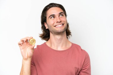 Beyaz arka planda izole edilmiş Bitcoin tutan yakışıklı genç adam gülümserken yukarı bakıyor.