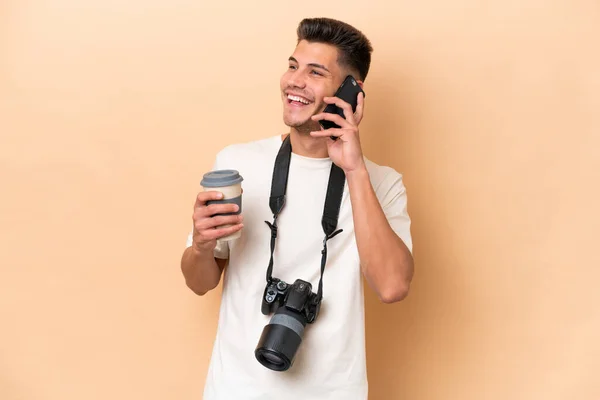 年轻的摄影师高加索人被隔离在米色背景下 手里拿着咖啡和手机 — 图库照片