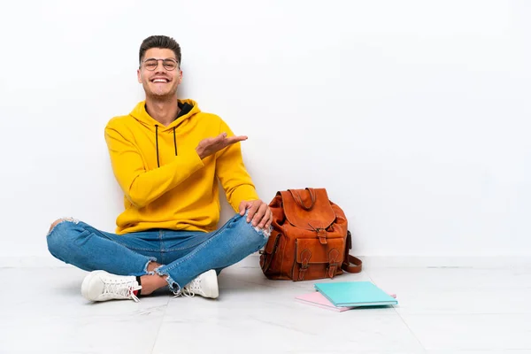 Young student caucasian man sitting one the floor isolated on white background presenting an idea while looking smiling towards