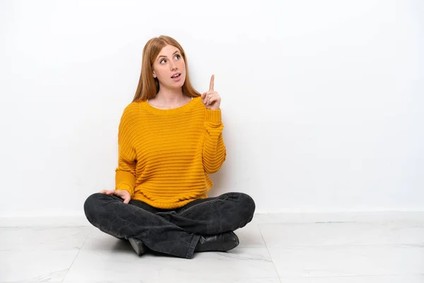 白い背景に隔離された床に座っている若い赤毛の女性は 指を指差すアイデアを考えます — ストック写真