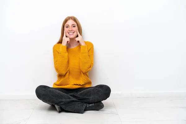 若い赤毛の女性が幸せと快適な表情で笑顔で白い背景に隔離された床に座って — ストック写真
