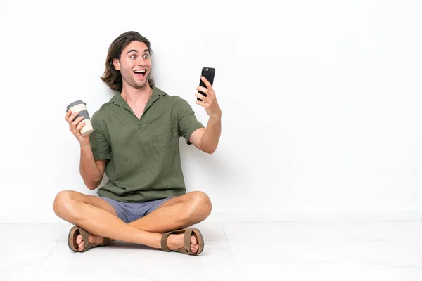 Mladý Pohledný Muž Sedí Podlaze Izolované Bílém Pozadí Drží Kávu — Stock fotografie