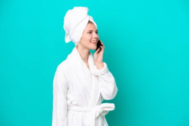 Bornozlu genç kadın mavi arka planda izole edilmiş cep telefonuyla sohbet ediyor.