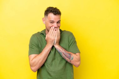 Sarı arka planda izole edilmiş beyaz, yakışıklı genç adam mutlu ve gülümseyen elleriyle ağzını kapatıyor.