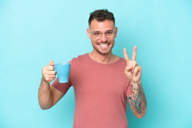Mavi arka planda izole edilmiş bir fincan kahve tutan genç beyaz adam gülümsüyor ve zafer işareti gösteriyor.