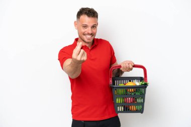 Beyaz arka planda izole edilmiş yiyecek dolu bir alışveriş sepeti tutan beyaz bir adam para hareketi yapıyor.