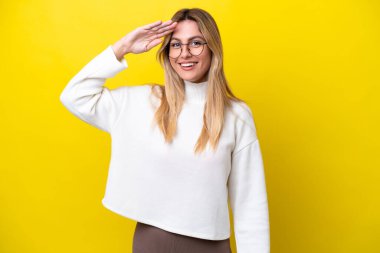 Young Uruguayan woman isolated on yellow background saluting with hand with happy expression