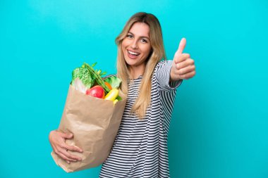 Uruguaylı genç bir kadın, elinde alışveriş poşeti ile mavi arka planda izole bir şekilde duruyor. Başparmakları havada çünkü iyi bir şey oldu.