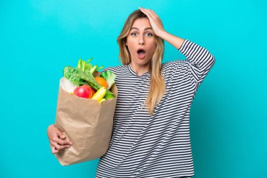 Genç Uruguaylı bir kadın elinde alışveriş çantası tutarken mavi arka planda sürpriz bir ifadeyle izole edilmiş.