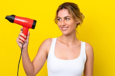Young Uruguayan woman holding a hairdryer isolate don yellow background with happy expression