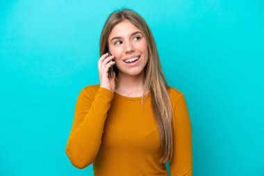 Mavi arka planda izole edilmiş genç beyaz bir kadın cep telefonuyla sohbet ediyor.