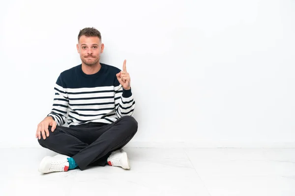 Mladý Muž Sedí Podlaze Izolovaný Bílém Pozadí Ukazuje Ukazováčkem Skvělý — Stock fotografie