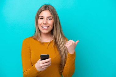 Genç beyaz kadın mavi arka planda izole edilmiş cep telefonu kullanıyor ve yan tarafı işaret ediyor.