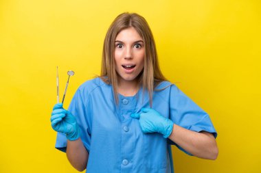 Dentist caucasian woman holding tools isolated on yellow background with surprise facial expression