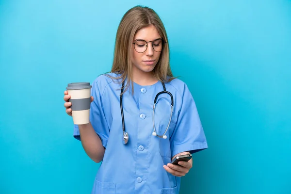 stock image Young surgeon doctor woman isolated on blue background holding coffee to take away and a mobile