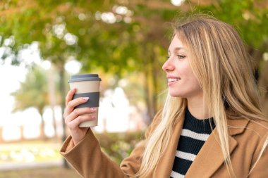 Genç, güzel, sarışın bir kadın elinde kahveyle dışarıda mutlu bir ifadeyle bekliyor.