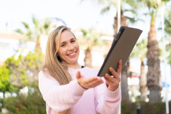 Young blonde woman holding a tablet at outdoors with happy expression