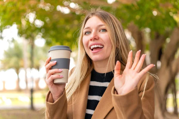 年轻漂亮的金发女子在户外端着一杯咖啡 带着愉快的表情向她致意 — 图库照片