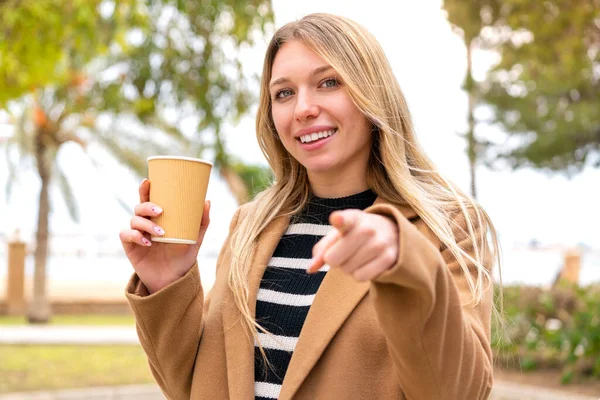 年轻漂亮的金发女人在户外端着咖啡 带着自信的表情指指点点你 — 图库照片