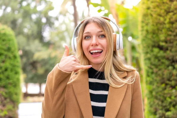 Молодая Симпатичная Блондинка Слушает Музыку Наушниках Улице Делает Телефонный Жест — стоковое фото