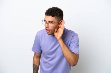 Young caucasian Brazilian man isolated on white background listening to something by putting hand on the ear