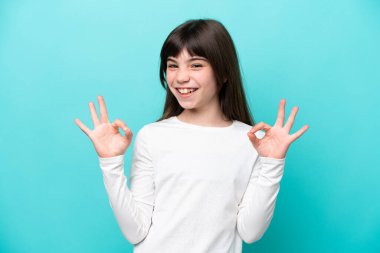 Mavi arka planda izole edilmiş küçük beyaz kız iki eliyle tamam işareti gösteriyor.