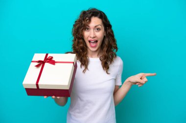 Elinde mavi arka planda izole edilmiş bir hediye tutan genç beyaz kadın şaşırdı ve parmağını yana çevirdi.
