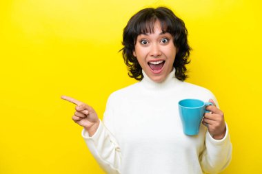 Arjantinli genç bir kadın elinde kahve fincanı tutarken sarı arka planda izole edilmiş şaşırmış ve parmağını kenarı işaret ediyor.