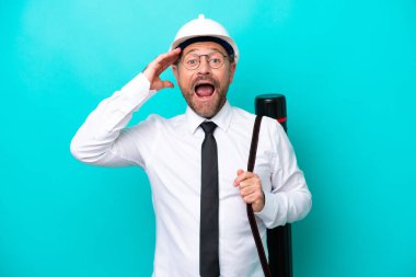 Miğferli orta yaşlı bir mimar elinde mavi arka planda, sürpriz bir ifadeyle izole edilmiş planlar tutuyor.