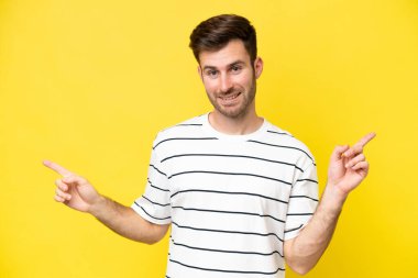 Sarı arka planda izole edilmiş genç beyaz adam parmak uçlarını işaret ediyor ve mutlu.