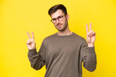 Sarı arka planda izole edilmiş genç beyaz adam iki eliyle zafer işareti gösteriyor.