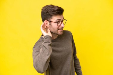 Sarı arka planda izole edilmiş genç beyaz adam elini kulağına koyarak bir şeyler dinliyor.