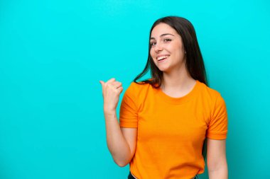 Mavi arka planda izole edilmiş genç beyaz kadın bir ürünü sunmak için kenarı işaret ediyor.