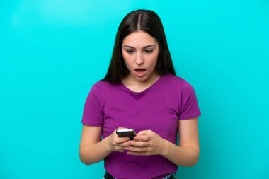 Young girl isolated on blue background looking at the camera while using the mobile with surprised expression