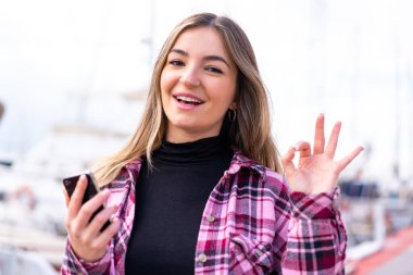 Genç, güzel Romen kadın açık havada cep telefonu kullanıyor ve tamam işareti yapıyor.