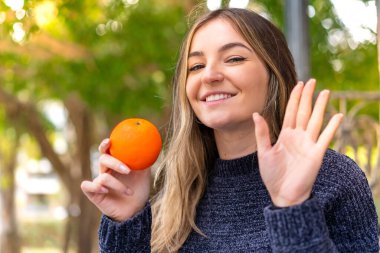 Genç ve güzel Romen kadın elinde bir portakalla elini sallayarak neşeli bir ifadeyle selam veriyor.