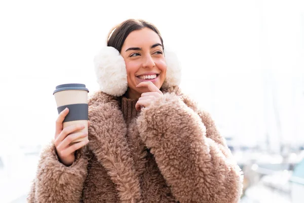 年轻女人穿着冬衣 抱着咖啡 站在室外 想着一个主意 就往旁边看 — 图库照片
