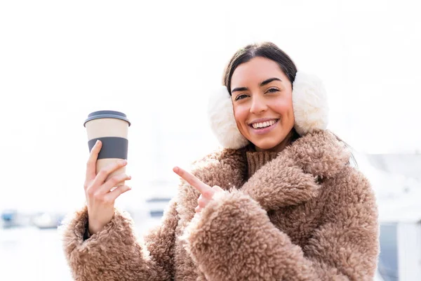 年轻女人穿着冬衣 拿着咖啡到户外 指着咖啡 — 图库照片