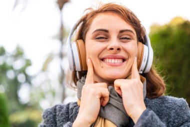 Açık havada müzik dinleyen esmer kadın mutlu ve hoş bir ifadeyle gülümsüyor.