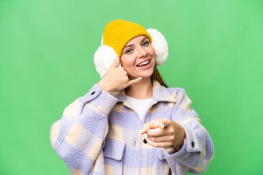 Sarışın genç bir kadın, izole edilmiş krom arka plan üzerinde kışlık manşetler takıyor telefon hareketleri yapıyor ve ön tarafı işaret ediyor.