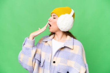 Sarışın genç bir kadın, izole edilmiş krom arka planda kışlık manşetler takıyor esniyor ve ağzını eliyle açık tutuyor.