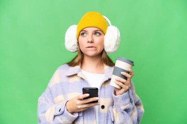 İzole edilmiş krom arka planda kışlık manşet takan genç sarışın kadın elinde kahve ve bir şeyler düşünürken bir cep telefonu tutuyor.