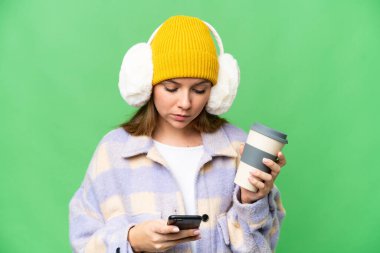 İzole edilmiş krom arka planda kışlık manşet takan genç sarışın bir kadın. Elinde kahve ve cep telefonu var.