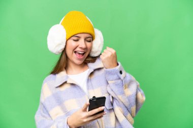 İzole edilmiş krom arka plan üzerinde kışlık manşet takan genç sarışın bir kadın. Telefon zafer pozisyonunda.