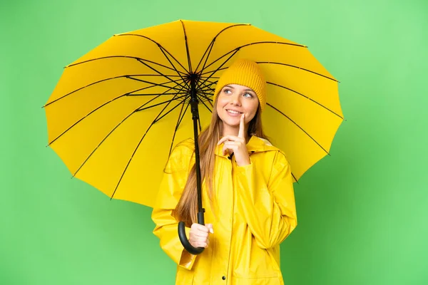若いですブロンド女性とともにレインプルーフコートと傘上の隔離されたクロマキー背景考えますAアイデアながら見上げます — ストック写真