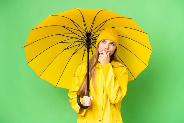 若いですブロンド女性とともにレインプルーフコートと傘上の隔離されたクロマキー背景持っています疑問 — ストック写真