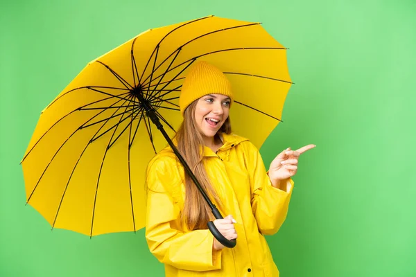 若いですブロンド女性とともにレインプルーフコートと傘上の隔離されたクロマキー背景指差し指で側面と製品を提示 — ストック写真