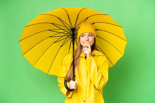 若いですブロンド女性とともにレインプルーフコートと傘上の隔離されたクロマキー背景持っています疑問と思考 — ストック写真